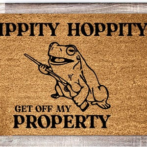 Może przedstawiać: Brązowa mata kokosowa z napisem "Hippity Hoppity Get Off My Property" i kreskówkową żabą trzymającą kij.
