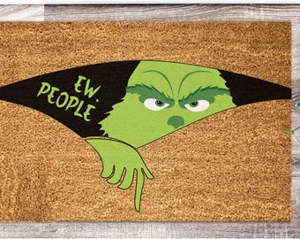 Puede incluir: Una alfombra rectangular con una superficie marrón texturizada. El diseño presenta un personaje verde de Grinch asomándose desde un recorte negro, con las palabras "EW. PEOPLE" en verde. La mano del Grinch apunta hacia abajo.