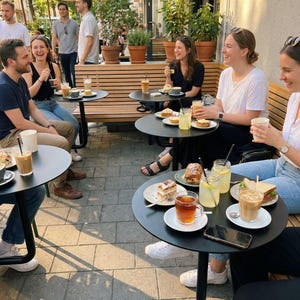 May include: Outdoor cafe scene with people seated at small black tables, enjoying drinks and food. Beverages include iced coffee, tea, and lemonade. Plates of pastries and sandwiches are visible. The setting is a sunny day with umbrellas overhead.