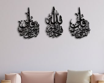 Puede incluir: Tres piezas de decoración de pared negras con caligrafía árabe en un ambiente de sala de estar moderna. Las piezas están montadas en una pared gris claro sobre un sofá rosa claro con un cojín blanco. La habitación tiene un suelo de madera clara y una mesa auxiliar de madera con un jarrón de plantas verdes.