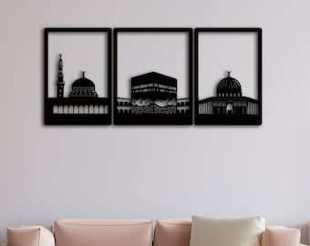 May include: Three black metal wall art pieces depicting the Kaaba, the Masjid al-Haram, and the Dome of the Rock. The art pieces are framed and hung on a white wall in a living room setting.