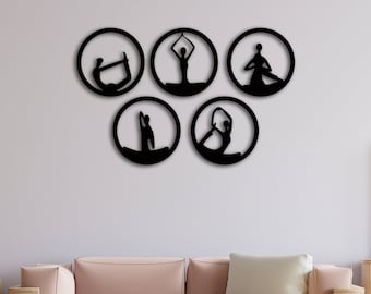 Puede incluir: Cinco piezas de decoración de pared de metal negro en forma de círculos con siluetas de personas haciendo poses de yoga. La decoración se muestra en una pared gris claro sobre un sofá rosa en una sala de estar.