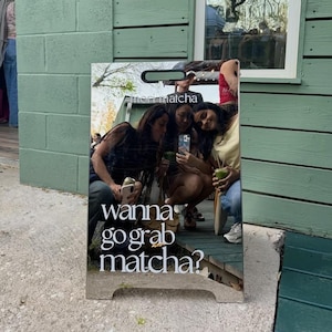 May include: A rectangular sign with the text "wanna go grab matcha?" in white. The sign reflects an image of three people holding drinks and a phone. The sign is outside a building with green siding and a brick wall.