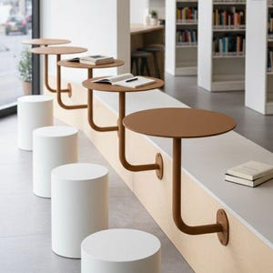 Op de afbeelding: Een modern bibliotheekinterieur met een rij kleine, ronde, bruine tafels die aan een lichtgekleurde muur zijn bevestigd. Witte cilindrische krukken staan voor de tafels. Boekenkasten vol boeken staan op de achtergrond.