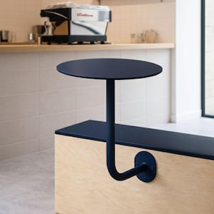 Puede incluir: Una mesa redonda azul marino fijada a un banco de madera. La mesa tiene una sola pata curva que se conecta al banco. El banco es de madera clara y tiene una parte superior azul marino. El fondo incluye una máquina de café y una estantería con plantas.