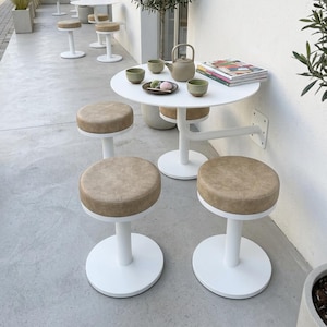 May include: Outdoor cafe setting with white round tables and stools with tan seats. A teapot, cups, and small pastries sit on a table. Olive trees and other greenery add to the ambiance. The scene is bright and airy.