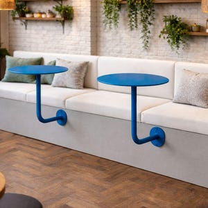 Op de afbeelding: Een modern café-interieur met een ingebouwde bank met witte kussens en een grijze basis. Twee kleine, ronde, blauwe tafels zijn aan de bank bevestigd. Decoratieve kussens in neutrale en groene tinten voegen kleur toe. Boven hangen geweven hanglampen.