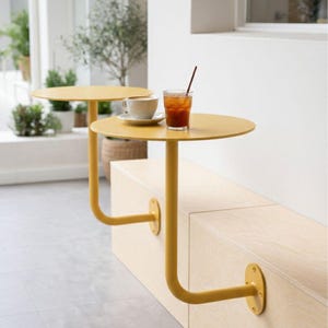 May include: Two yellow, round cafe tables attached to a light wood bench. One table holds a white teacup and saucer, the other has a glass of iced tea with a straw. The tables have a unique, curved leg design.