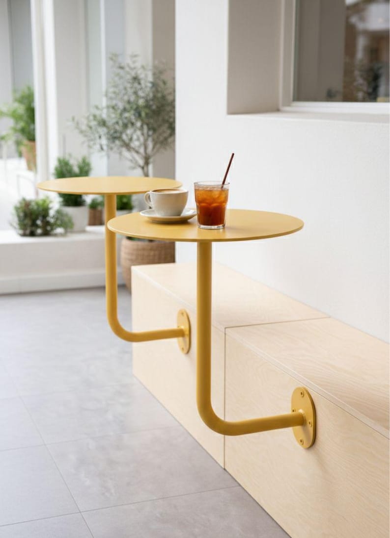 May include: Two yellow, round cafe tables attached to a light wood bench. One table holds a white teacup and saucer, the other has a glass of iced tea with a straw. The tables have a unique, curved leg design.