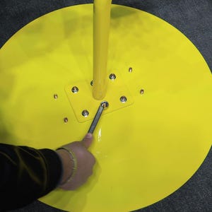 May include: Bright yellow, circular base with a curved, vertical yellow pole. A person is using a wrench to tighten a bolt on the base. The base has a square metal plate with multiple bolts. The background is a dark gray surface.