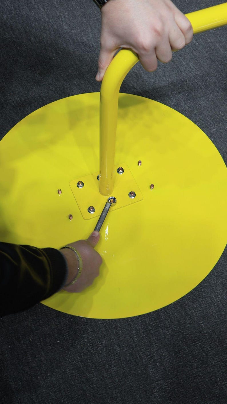 May include: Bright yellow, circular base with a curved, vertical yellow pole. A person is using a wrench to tighten a bolt on the base. The base has a square metal plate with multiple bolts. The background is a dark gray surface.