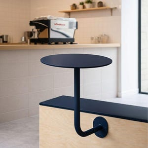 May include: A navy blue, round table attached to a wooden bench. The table is supported by a curved, navy blue metal pole. The bench is made of light-colored wood and has a navy blue top. The background includes a coffee machine and a shelf with plants.