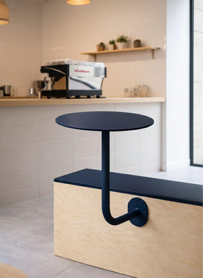 May include: A navy blue, round table attached to a wooden bench. The table is supported by a curved, navy blue metal pole. The bench is made of light-colored wood and has a navy blue top. The background includes a coffee machine and a shelf with plants.