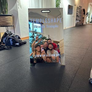 May include: A silver sign with the text "TEMPLESHAPE Health Temple" and the slogan "SHOW UP & GLOW UP!"  The sign is reflecting a group of people smiling and taking a selfie.