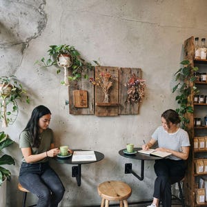 May include: Two people are seated at small, black tables in a cafe. One person is writing in a notebook, and the other is holding a cup of green beverage. The cafe has a rustic aesthetic with wooden shelves and wall decor.