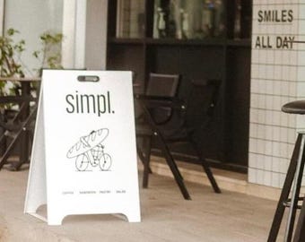 Puede incluir: Un letrero blanco en forma de A con la palabra "simpl." en texto negro. El letrero tiene una ilustración en blanco y negro de una persona montando en bicicleta con una baguette a la espalda. El letrero también tiene el texto "Coffee, Sandwich, Pastry, Salad" en texto negro pequeño.