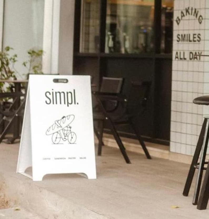May include: A white A-frame sign with the word "simpl." in black text. The sign has a black and white illustration of a person riding a bicycle with a baguette on their back. The sign also has the text "Coffee, Sandwich, Pastry, Salad" in small black text.