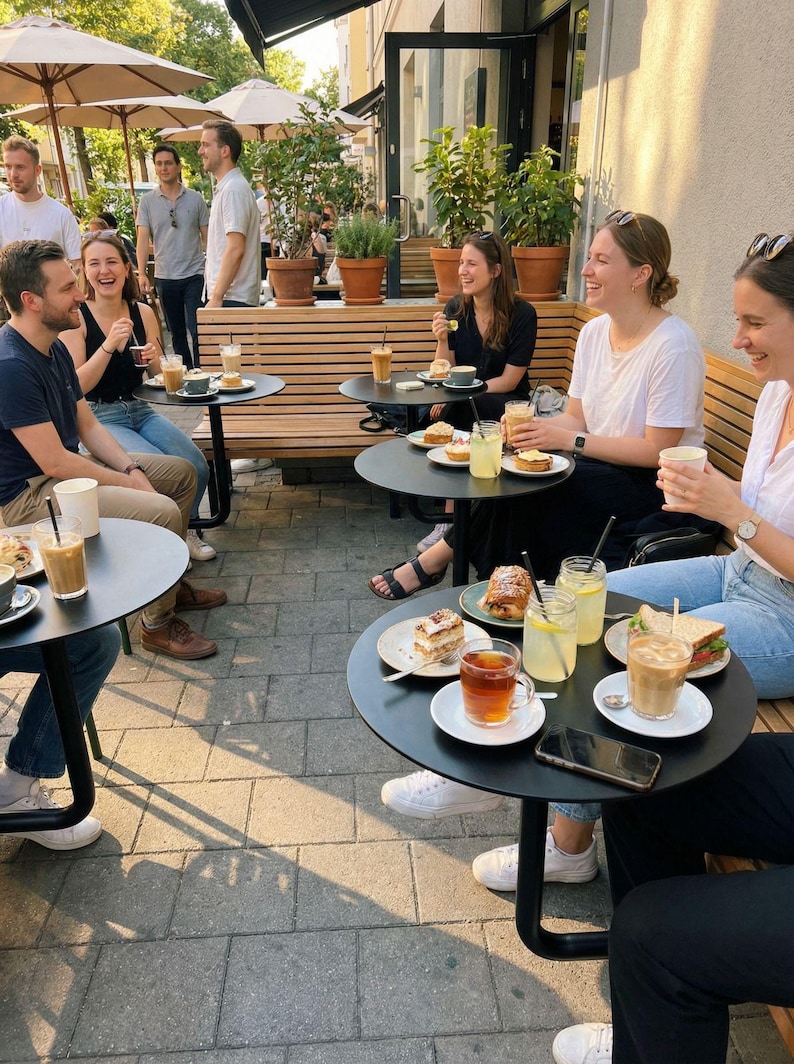 May include: Outdoor cafe scene with people seated at small black tables, enjoying drinks and food. Beverages include iced coffee, tea, and lemonade. Plates of pastries and sandwiches are visible. The setting is a sunny day with umbrellas overhead.