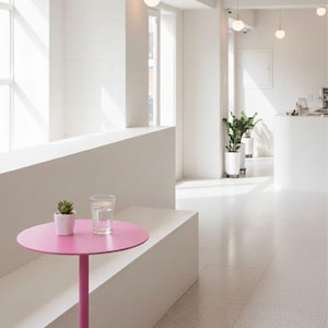 May include: A modern cafe interior with a bright, minimalist design. A small, round, pink table is attached to a white bench. A glass of water and a small potted plant sit on the table. Overhead, spherical pendant lights hang from the ceiling.