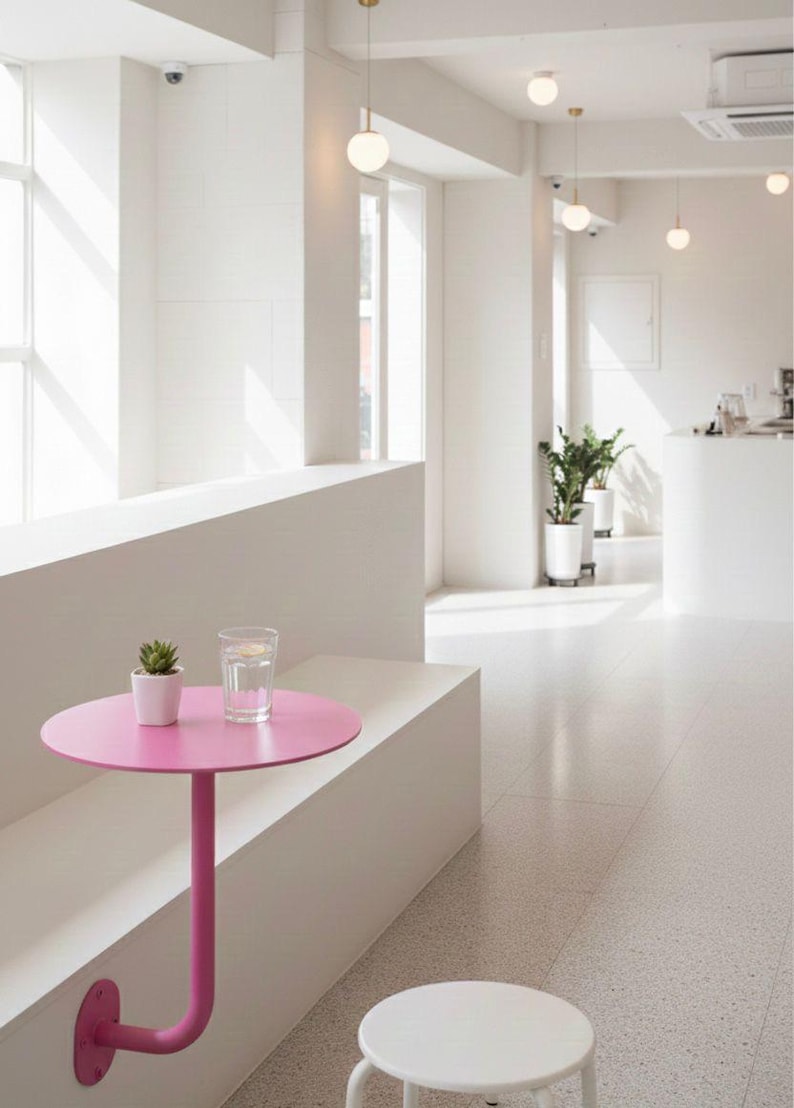 May include: A modern cafe interior with a bright, minimalist design. A small, round, pink table is attached to a white bench. A glass of water and a small potted plant sit on the table. Overhead, spherical pendant lights hang from the ceiling.