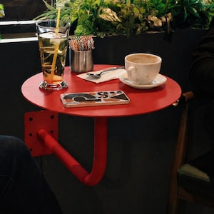 May include: A small, round, red table with a curved support arm is attached to a dark wall. A glass of iced tea, a coffee cup, and a smartphone rest on the table. Green plants are in the background.