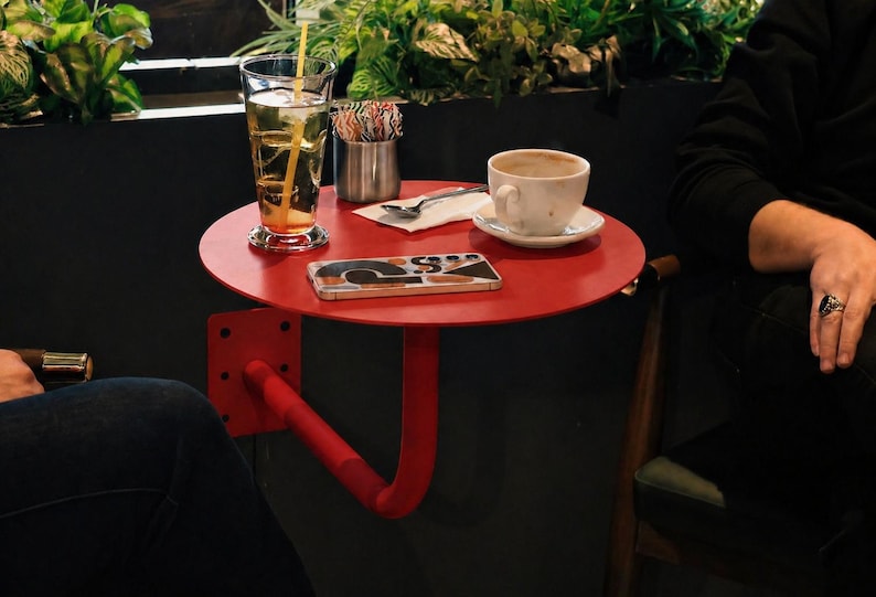 May include: A small, round, red table with a curved support arm is attached to a dark wall. A glass of iced tea, a coffee cup, and a smartphone rest on the table. Green plants are in the background.