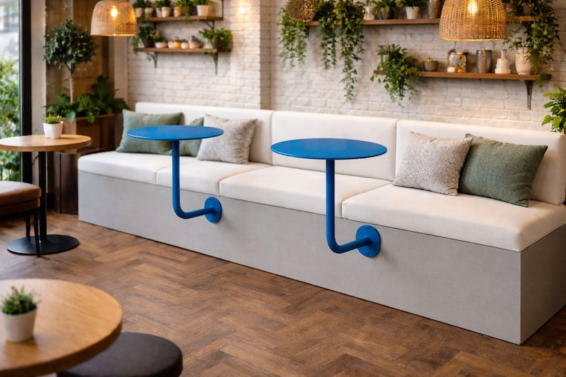 May include: A modern cafe interior featuring a built-in bench with white cushions and gray base. Two small, round, blue tables are attached to the bench. Decorative pillows in neutral and green tones add a pop of color. The space is decorated with plants.