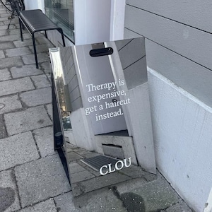 Op de afbeelding: Een glanzend zilveren bord met de tekst "Therapy is expensive, get a haircut instead." en de naam "CLOU". Het bord staat buiten en leunt tegen een gebouw.