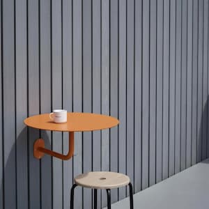 May include: An orange, wall-mounted, round table with a curved support arm. A white mug sits on the table. A wooden stool with black legs is in front of the table. The background is a gray, vertically-lined wall.