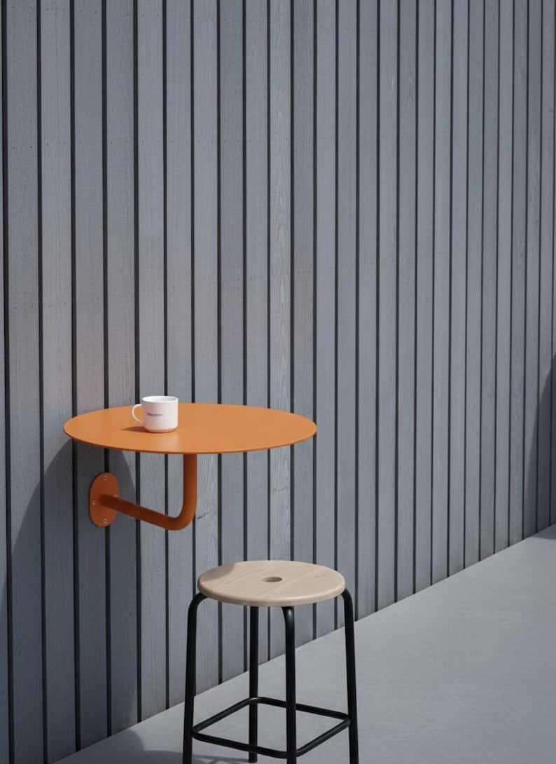 May include: An orange, wall-mounted, round table with a curved support arm. A white mug sits on the table. A wooden stool with black legs is in front of the table. The background is a gray, vertically-lined wall.