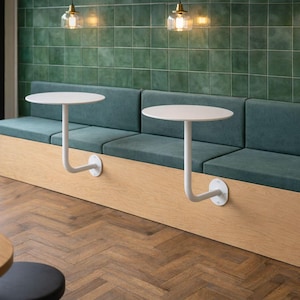 May include: A modern cafe seating area featuring a built-in bench with teal cushions and a light wood base. Two small, round, white tables are attached to the bench with curved metal supports. The background includes a green tiled wall and large windows.