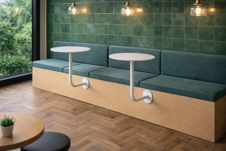 May include: A modern cafe seating area featuring a built-in bench with teal cushions and a light wood base. Two small, round, white tables are attached to the bench with curved metal supports. The background includes a green tiled wall and large windows.