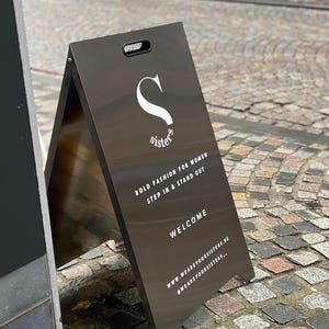 May include: A black A-frame sign with white text that reads "Sisters" and "Bold fashion for women step in & stand out". The sign also includes the text "Welcome" and the website address "www.wearyoursisters.nl" and the hashtag "#wearyoursisters..."