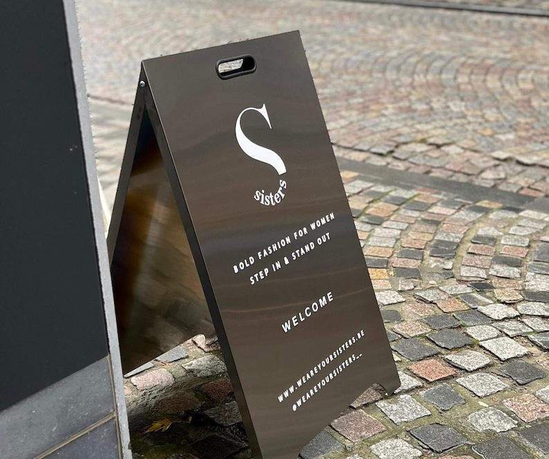 May include: A black A-frame sign with white text that reads "Sisters" and "Bold fashion for women step in & stand out". The sign also includes the text "Welcome" and the website address "www.wearyoursisters.nl" and the hashtag "#wearyoursisters..."