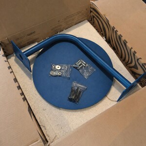 May include: A blue metal table leg and a round blue tabletop are inside a cardboard box. The leg is curved with flat mounting plates on each end. Small hardware bags with screws and washers are also included.