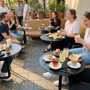 May include: Outdoor cafe scene with people seated at black tables, enjoying drinks and food. Beverages include iced coffee, tea, and lemonade. Food items include pastries and sandwiches. The setting is a sunny day with umbrellas and potted plants.