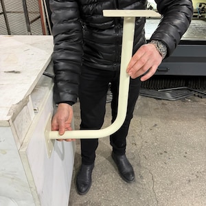May include: A cream-colored, L-shaped bracket with a flat top and a curved bottom section. The bracket is held by a person, showcasing its design and potential use in construction or furniture. The bracket is made of a solid material.