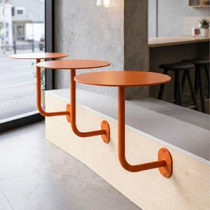 May include: Three orange, round tables attached to a curved wooden bench. The tables have a unique design with a single curved leg that attaches to the bench. The tables are a vibrant orange color, and the bench is made of light-colored wood.
