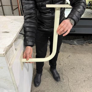 May include: A cream-colored, J-shaped bracket with a flat top and a curved bottom section. The bracket is held by a person. The bracket is designed to be mounted on a surface.