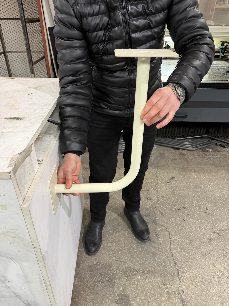 May include: A cream-colored, J-shaped bracket with a flat top and a curved bottom section. The bracket is held by a person. The bracket is designed to be mounted on a surface.