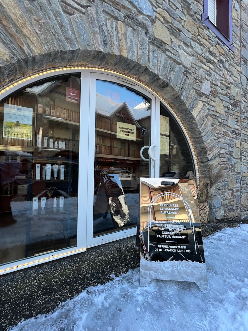 May include: A silver sign with black text advertising a massage chair experience. The sign reads "Discover massage differently. Treat yourself to our new massage chair experience. Offer yourself 20 minutes of absolute relaxation. EIMA."