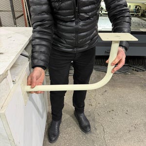 May include: A cream-colored, L-shaped bracket with a square mounting plate and a curved support arm. The bracket is held by a person wearing a black jacket and pants. The bracket appears to be made of metal.
