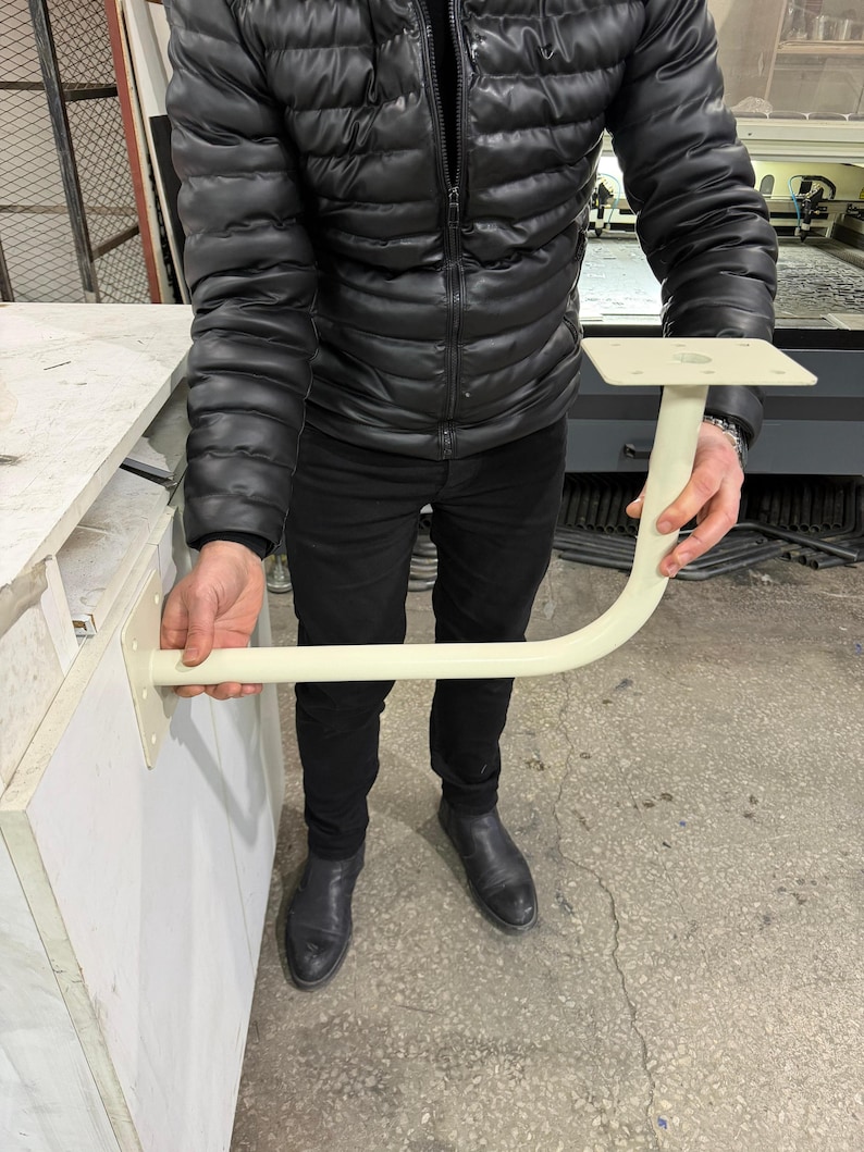 May include: A cream-colored, L-shaped bracket with a square mounting plate and a curved support arm. The bracket is held by a person wearing a black jacket and pants. The bracket appears to be made of metal.