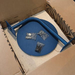 May include: A blue metal table leg and a blue circular tabletop are inside a cardboard box. The table leg is curved and has mounting plates on each end. Several small bags of hardware are also included.