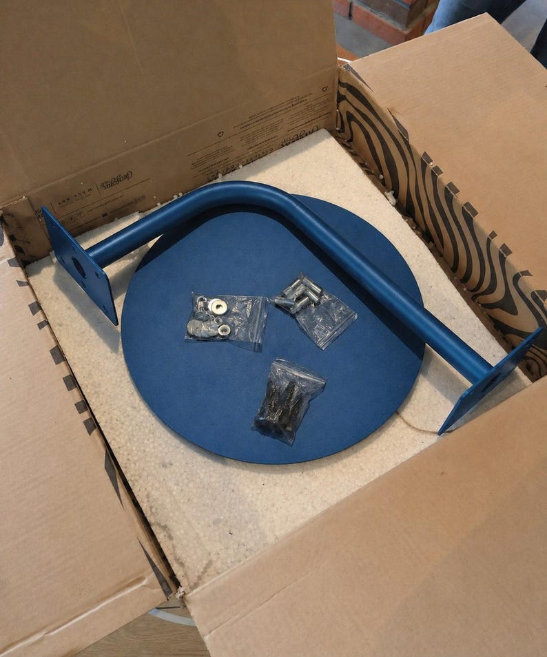 May include: A blue metal table leg and a blue circular tabletop are inside a cardboard box. The table leg is curved and has mounting plates on each end. Several small bags of hardware are also included.