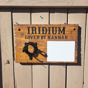 May include: Wooden sign with the word "IRIDIUM" and "LOVED BY HANNAH" in black lettering. The sign has a black horseshoe-shaped object attached and a white rectangular space. The sign is mounted on a wooden door.