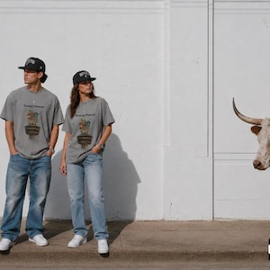 May include: A grey short-sleeve t-shirt featuring a graphic of a potted plant with the text "Flower Power" above it, paired with blue denim jeans and a black baseball cap. The design includes a small illustration of a brown animal in a pot.