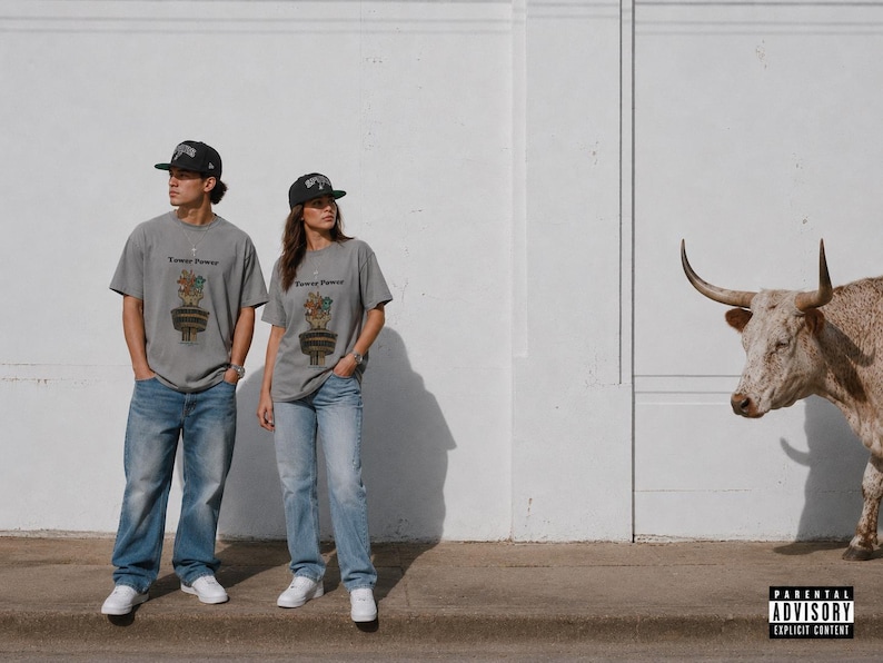 May include: A grey short-sleeve t-shirt featuring a graphic of a potted plant with the text "Flower Power" above it, paired with blue denim jeans and a black baseball cap. The design includes a small illustration of a brown animal in a pot.