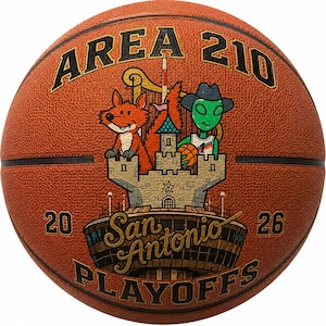 May include: A textured orange basketball featuring the text AREA 210 at the top and 20 26 on the sides. The center graphic depicts a cartoon fox and a green alien wearing a cowboy hat behind a stone castle, with San Antonio PLAYOFFS below.