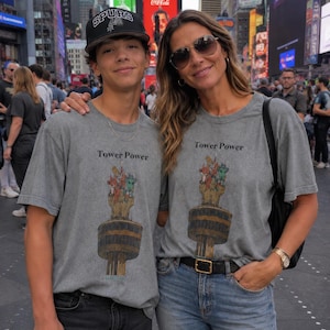 May include: Two grey crew-neck t-shirts featuring a graphic print of a tall tower with cartoon characters at the top and the text Tower Power. The shirts are worn with black jeans, a black baseball cap, blue jeans, and a black leather belt.